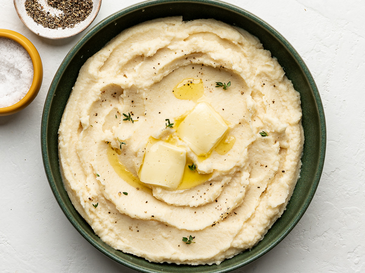Celeriac mash in green bowl topped with butter with a serving spoon and pinch bowls of salt and pepper on the side.