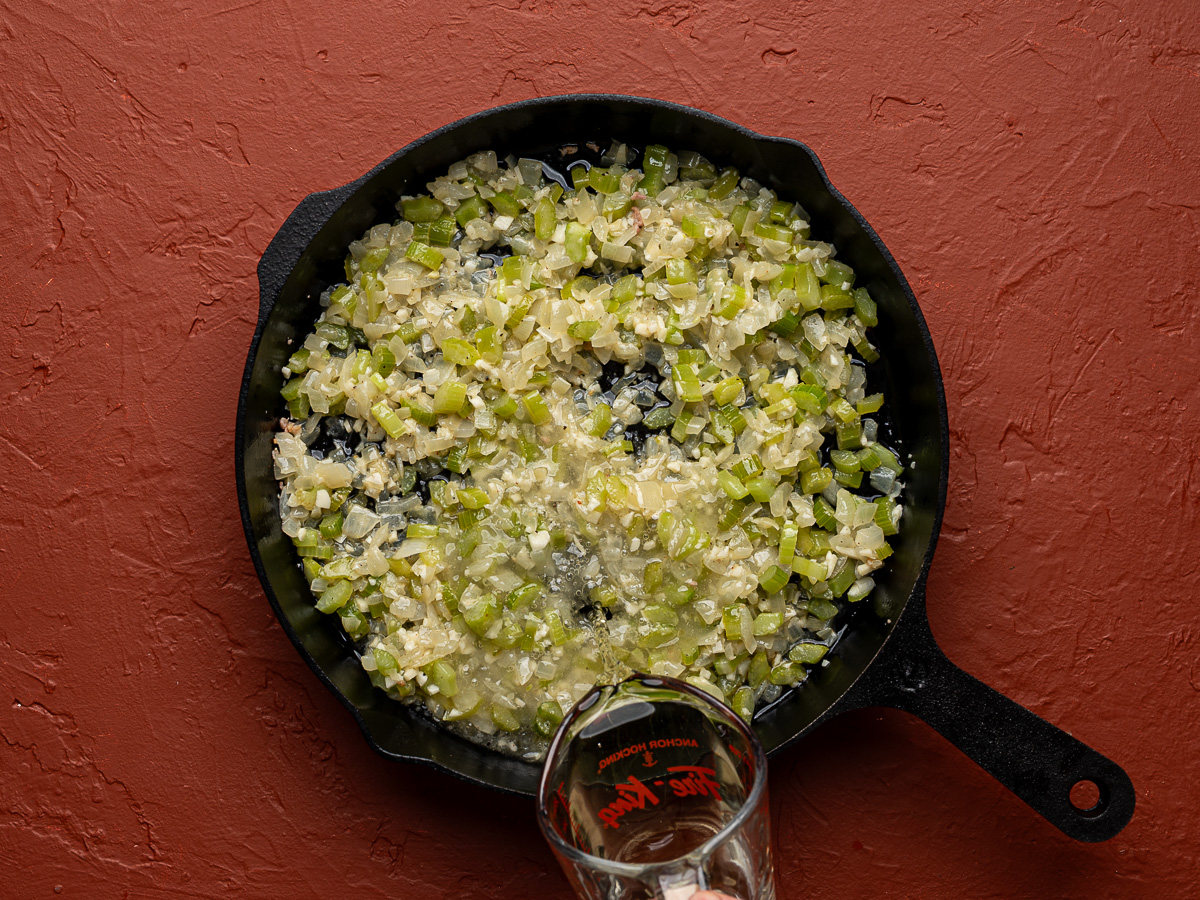 White wine getting poured over sautéed onion, celery and garlic in black skillet.