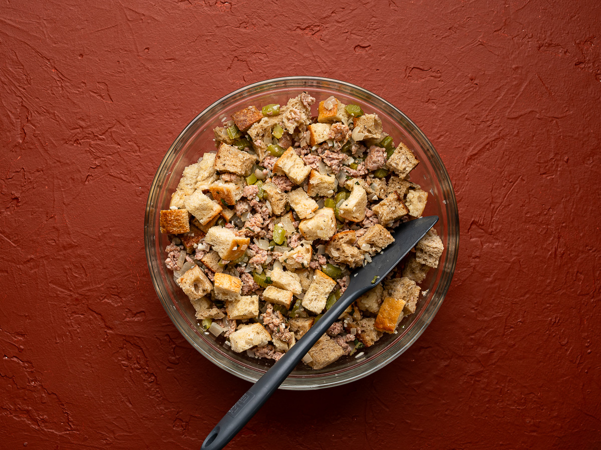 Bread cubes tossed with sausage mixture in glass bowl.