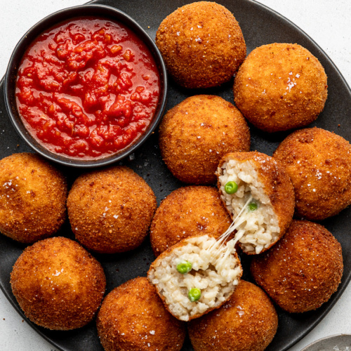 Fried arancini balls on a black plate with a bowl of marinara sauce on the side.