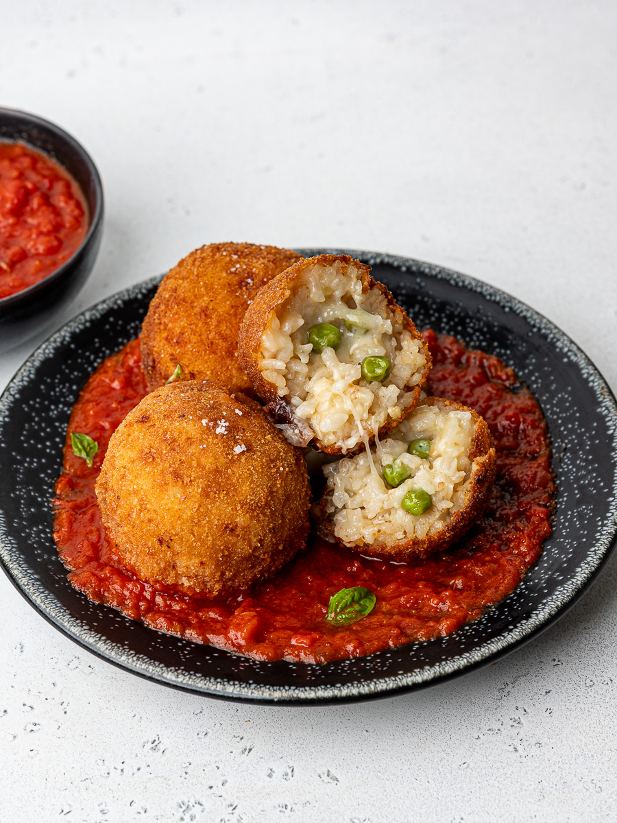 Three arancini balls sitting on marinara sauce on a black plate with one arancini cut open.