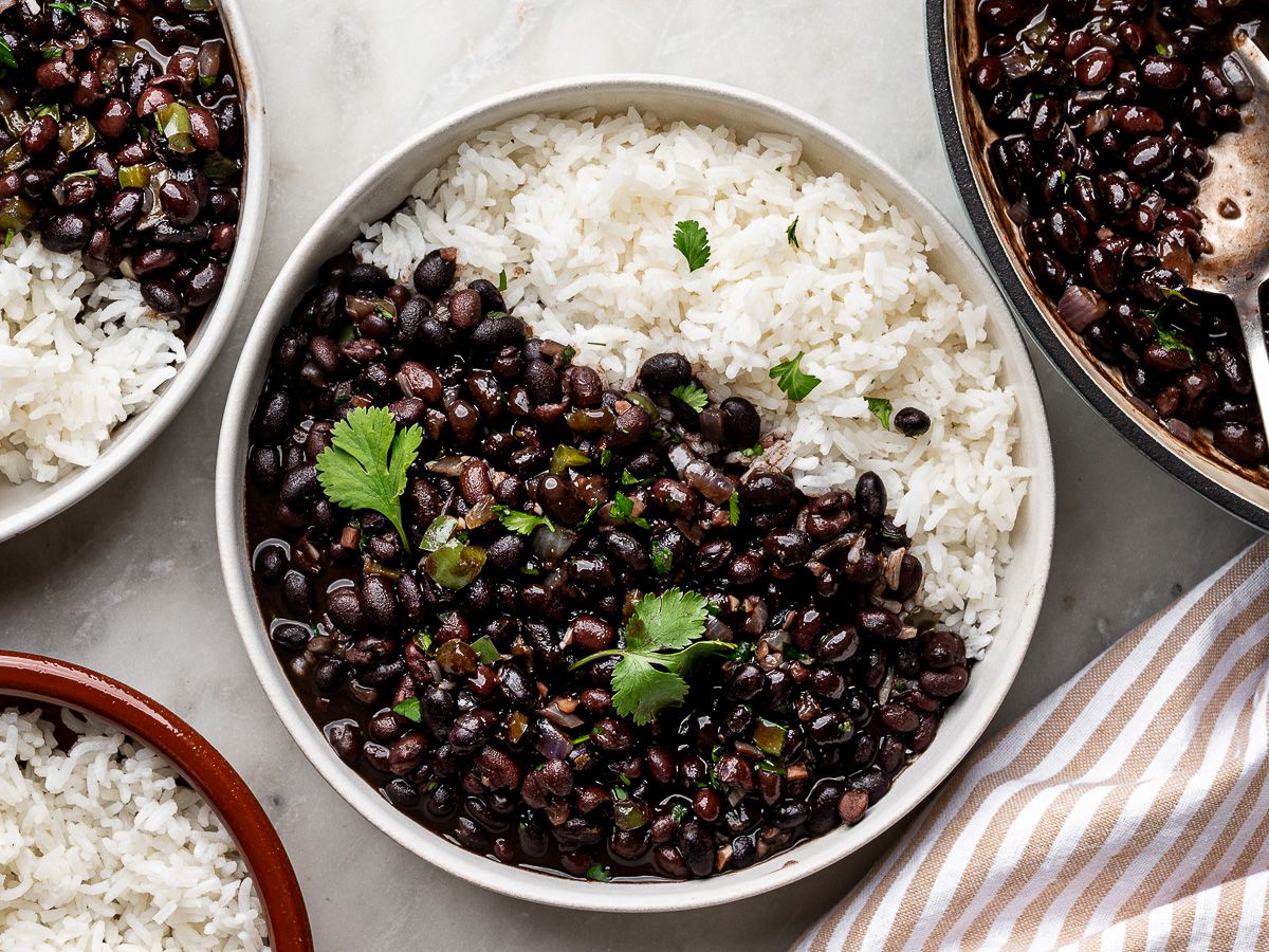 Cooked canned black beans garnished with cilantro served over white rice in a white bowl, with the skillet of beans on the side as well as a separate bowl of plain rice.