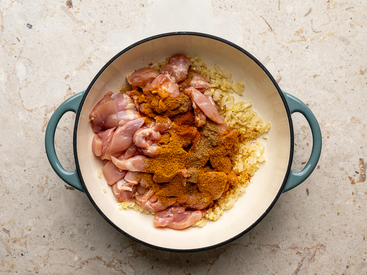 Raw chicken pieces added to the pan of sautéed aromatics with curry powder, garam masala, and cayenne, starting to absorb the spices.