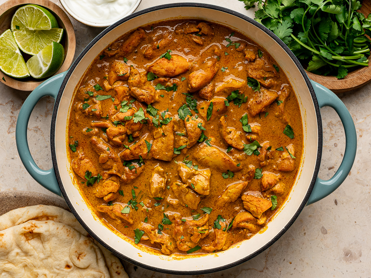 Finished coconut chicken curry in a pan, topped with fresh cilantro shown with naan, lime wedges and yogurt on the side.