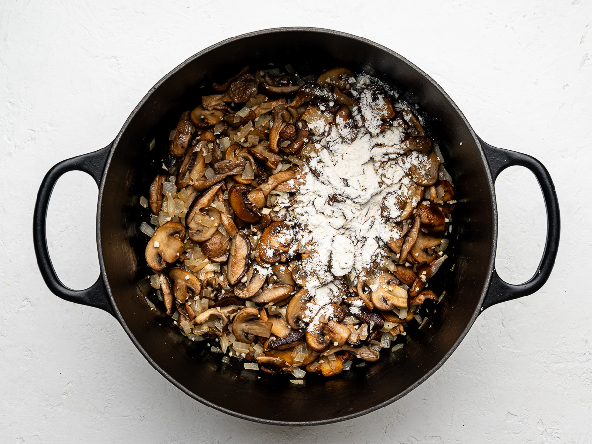 Flour and Dijon mustard being stirred into mushrooms and onions in a pot.