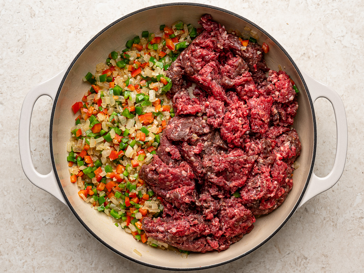 Ground beef and aromatics getting cooked in white skillet.