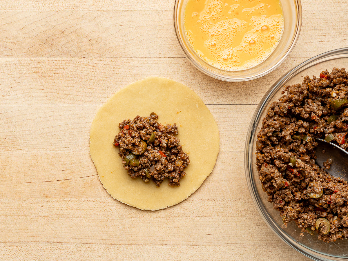 Filling an empanada disc with beef mixture and bowl of egg wash on the side.