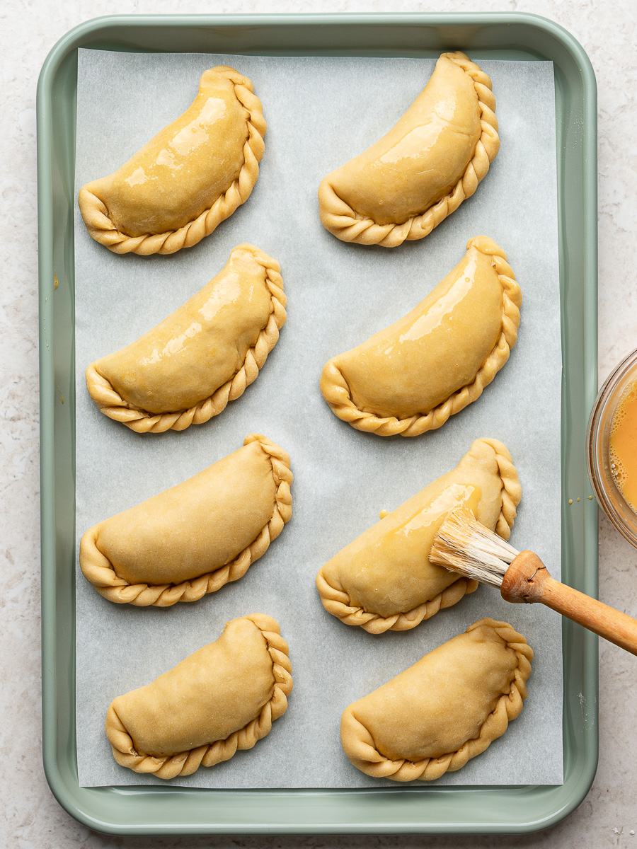 Brushing egg wash on unbaked empanadas with rope seal on baking sheet.