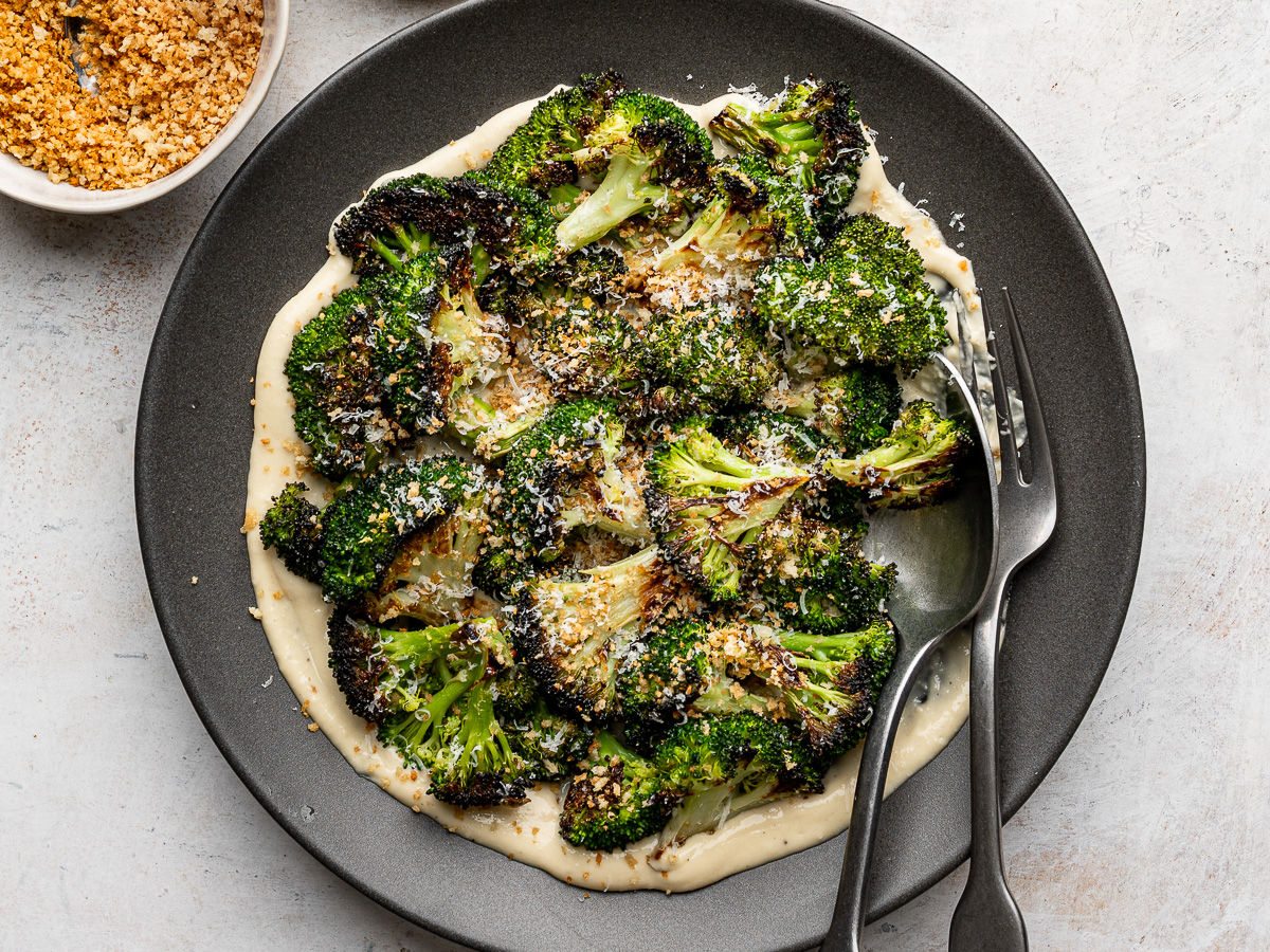 Close up of plated roasted broccoli florets arranged over a layer of Caesar dressing, sprinkled with toasted breadcrumbs and freshly grated Parmesan.
