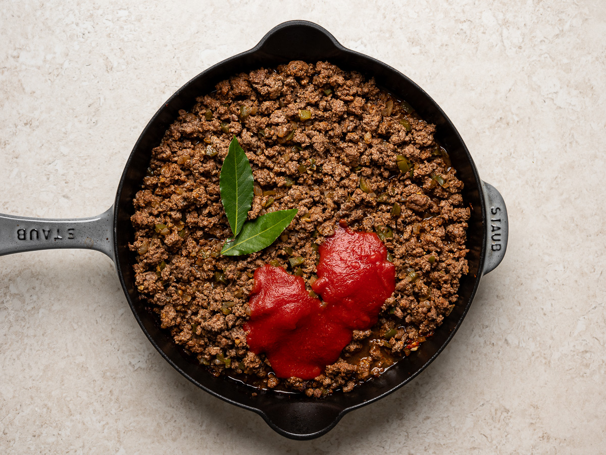 Adding tomato sauce and bay leaves to browned ground beef in skillet.
