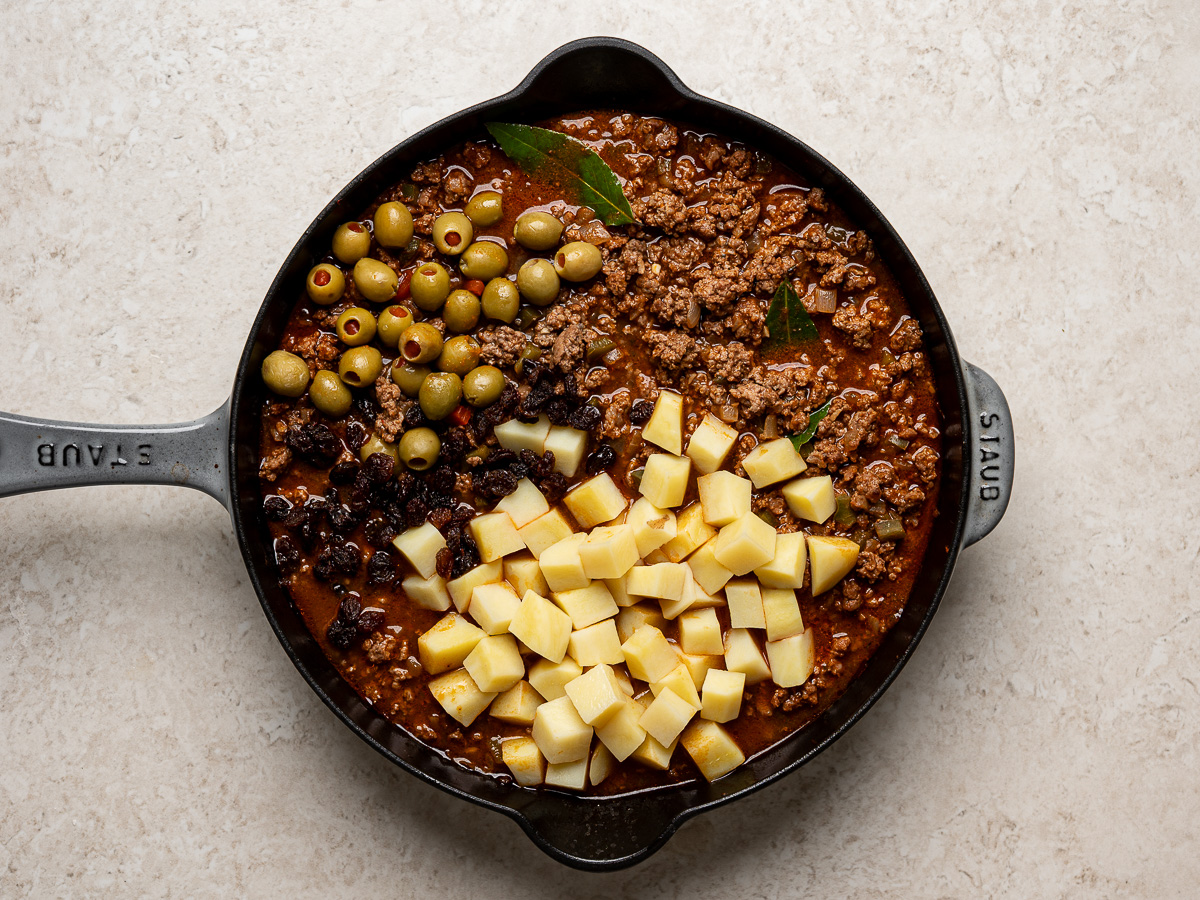 Adding par-boiled potatoes, olives, and raisins to browned ground beef mixture in skillet.