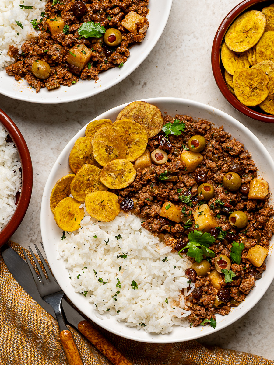 Two bowls of Cuban Picadillo served over white rice with plantain chips on the side.