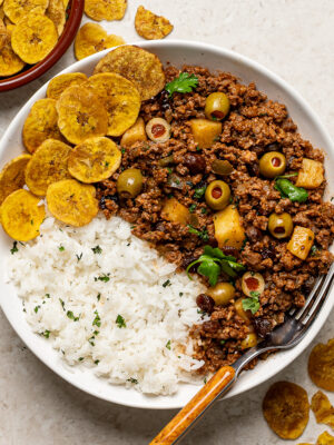 Cuban Picadillo served over white rice in a white bowl with plantain chips on the side.
