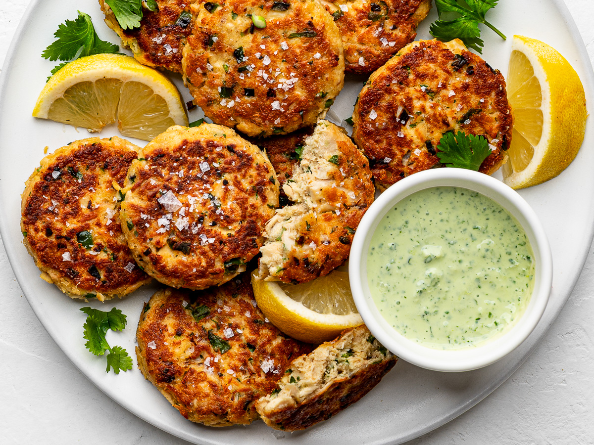 Close-up of pan-fried tuna patties on white plate with lemon wedges and dipping sauce on the side.