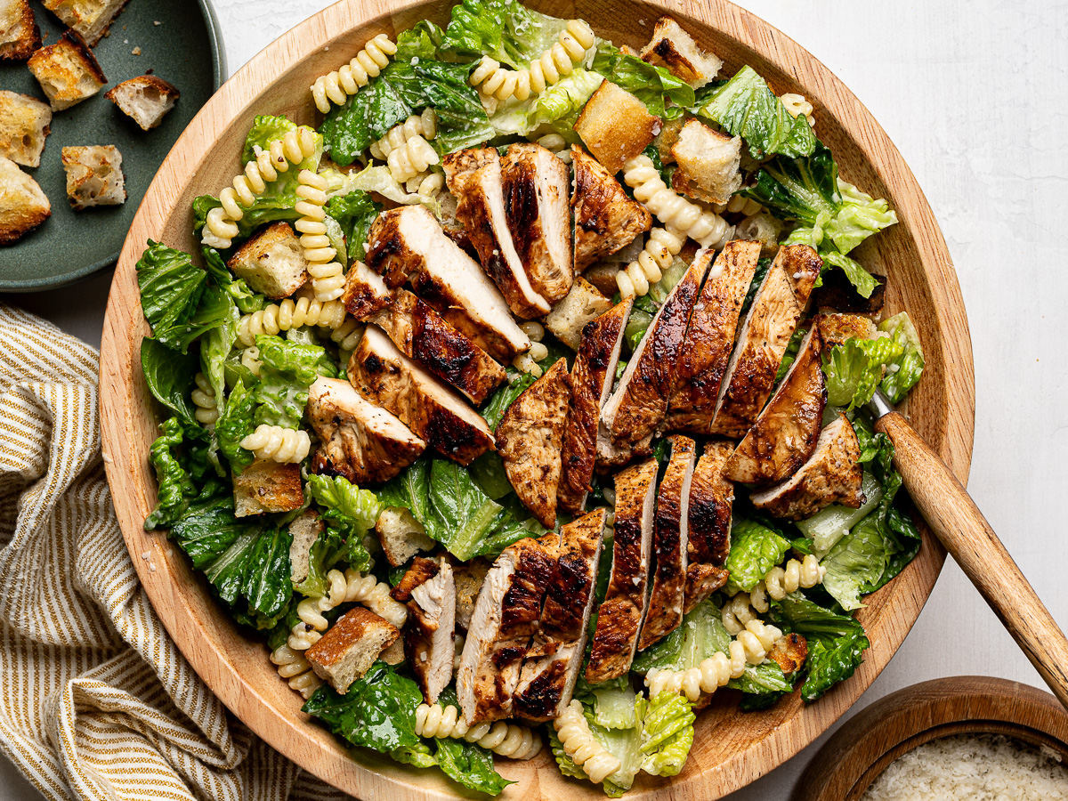 Caesar pasta salad in wooden bowl topped with chicken slices and croutons.