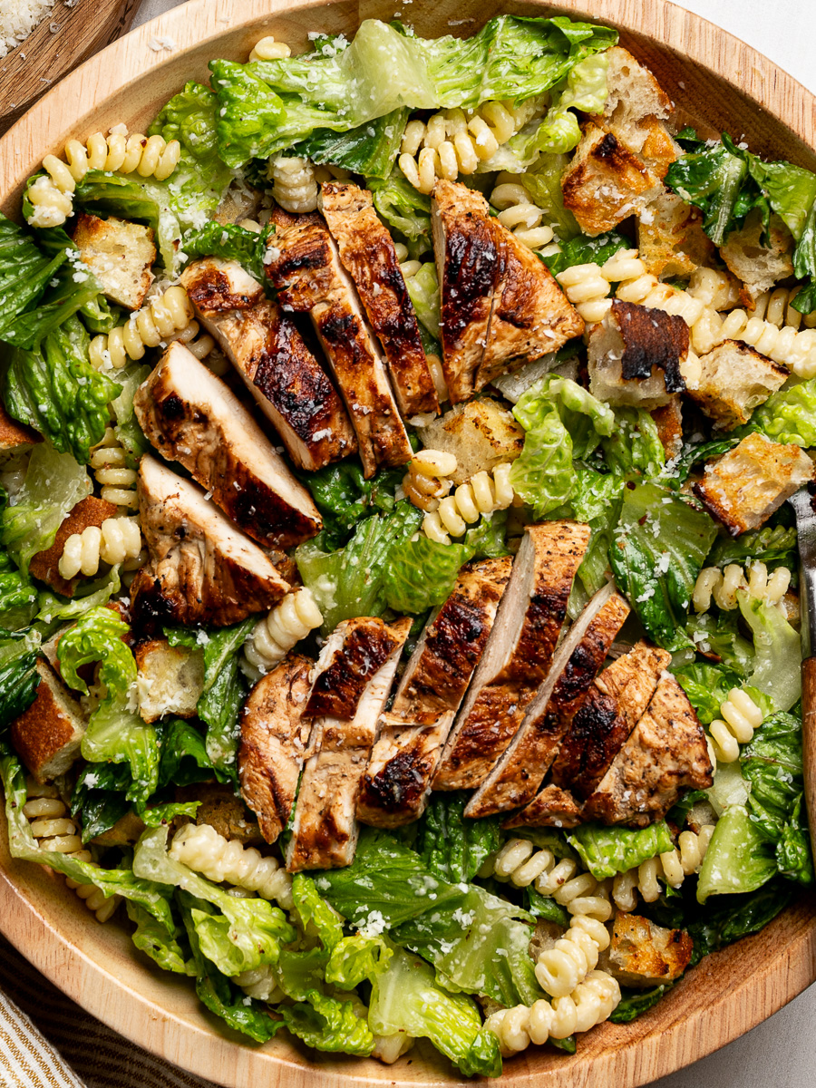 Close-up of Caesar pasta salad in wooden bowl topped with chicken slices and croutons.