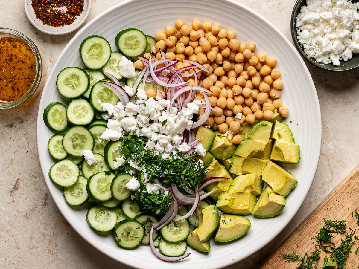 Sliced cucumbers, chopped avocados, chickpeas, sliced red onion, crumbled feta, and chopped herbs added to a large white serving bowl.