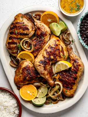 Grilled mojo chicken leg quarters on a white platter with sautéed onion rings and bowls of black beans and rice on the side.