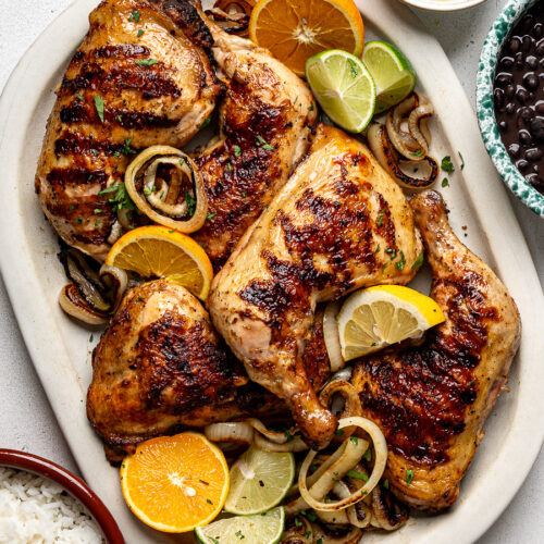 Grilled mojo chicken leg quarters on a white platter with sautéed onion rings and bowls of black beans and rice on the side.