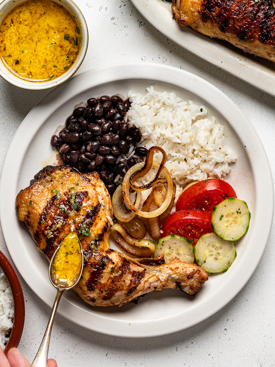 Drizzling mojo sauce over grilled mojo chicken leg served on a white plate with rice, beans, sliced tomatoes and cucumbers on the side.