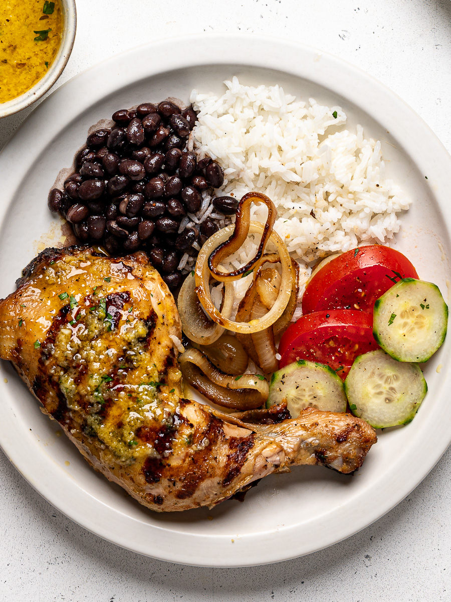 Grilled mojo chicken served on a white plate with rice, beans, sliced tomatoes and cucumbers on the side.