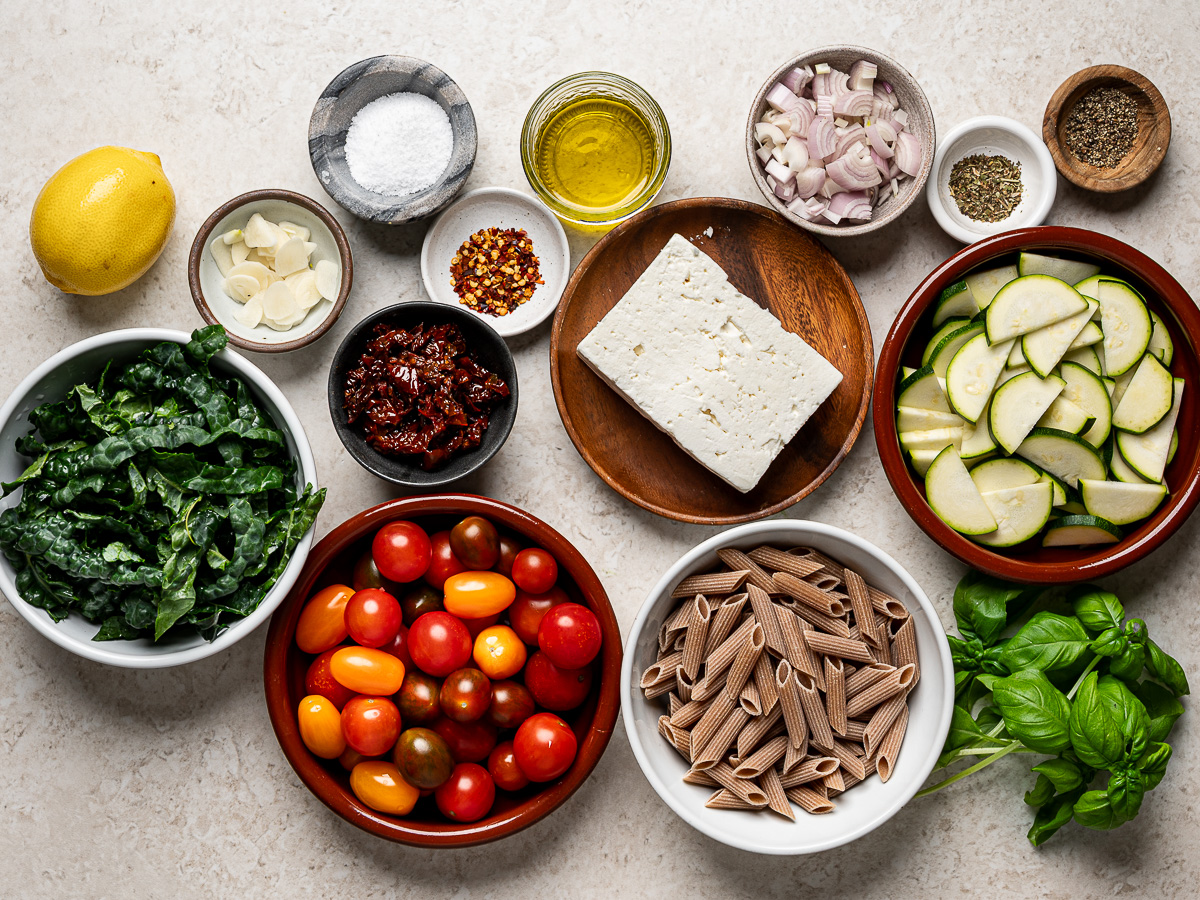 Recipe ingredients—dried penne pasta, cherry tomatoes, kale, zucchini, sun-dried tomatoes, block of feta, diced shallots, fresh basil, and spices— prepared in individual bowls.