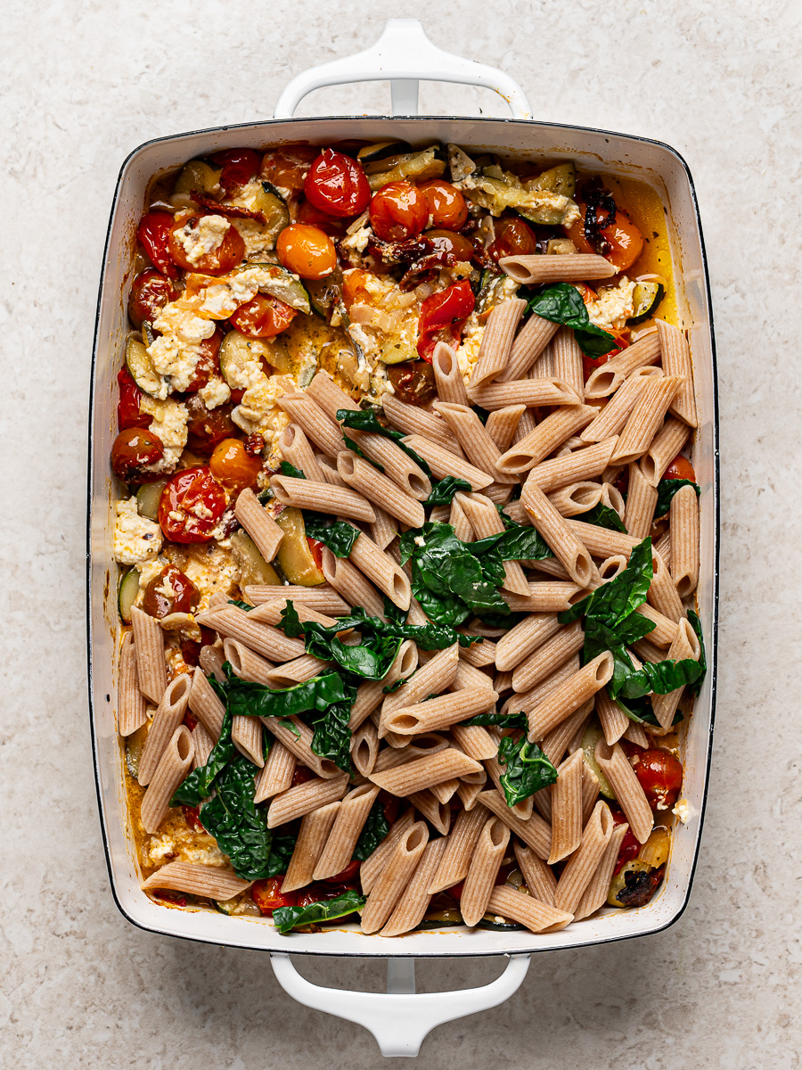 Adding cooked penne pasta and kale to the baked feta and vegetable mixture in baking dish.