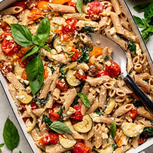 Close-up of baked feta, cherry tomatoes, penne pasta and zucchini in baking dish garnished with fresh basil leaves.