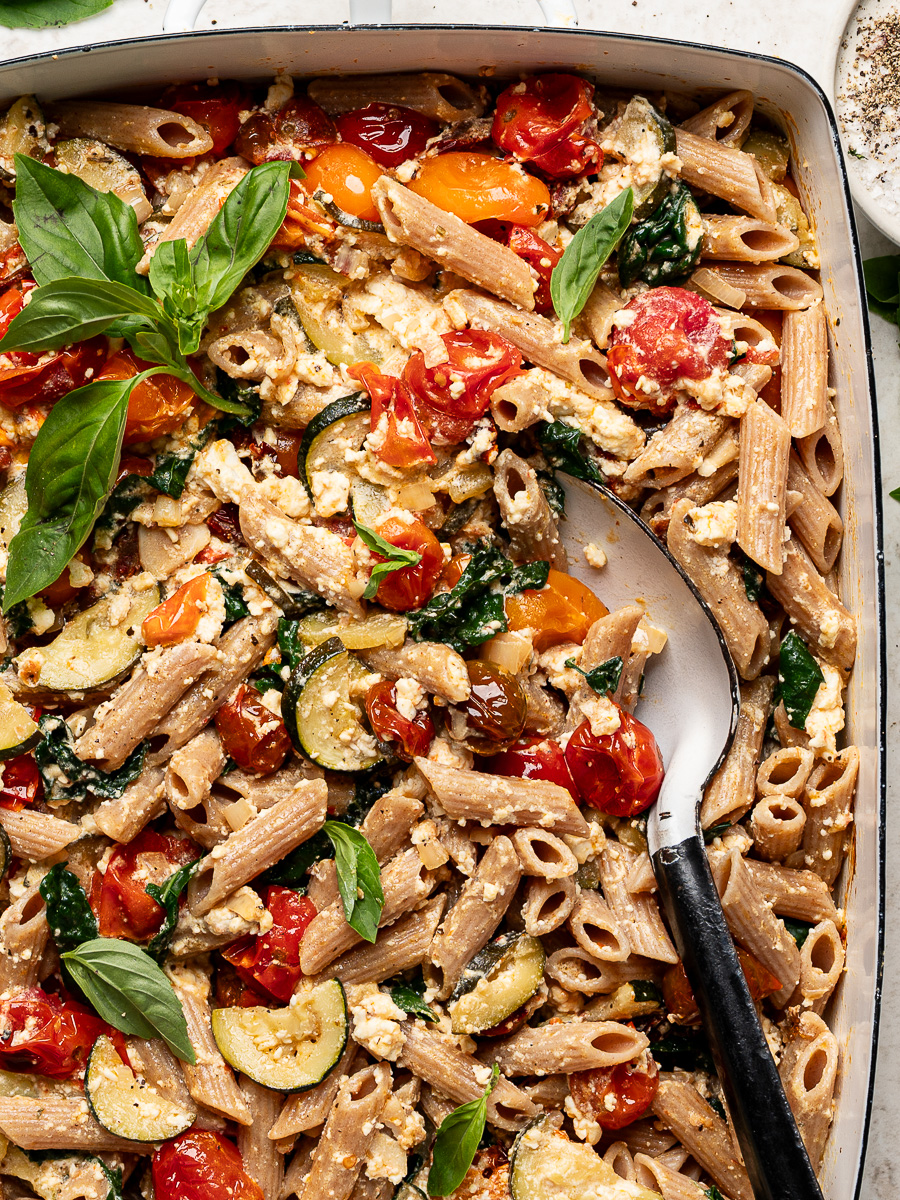 Close-up of baked feta, cherry tomatoes, penne pasta and zucchini in baking dish garnished with fresh basil leaves.