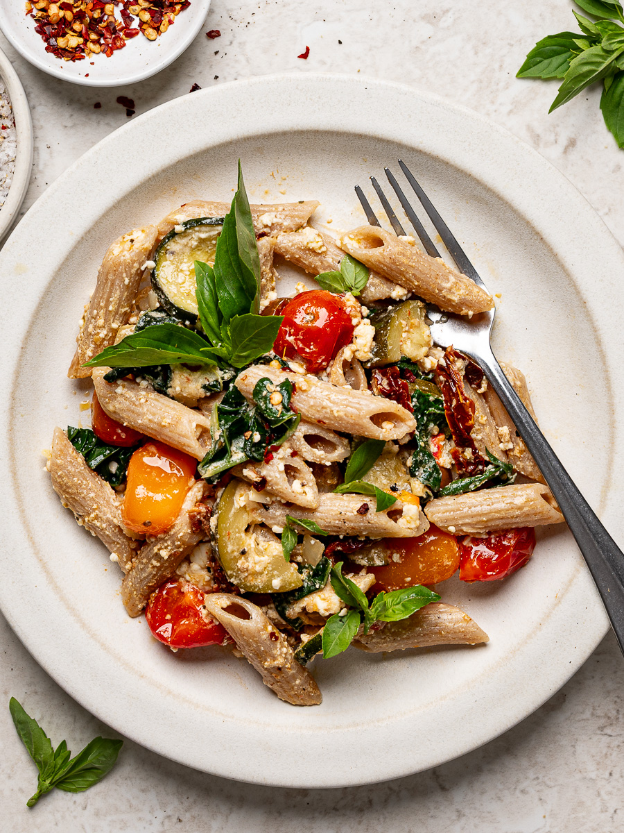 Baked feta pasta with burst tomatoes and zucchini served on a white plate garnished with fresh basil.