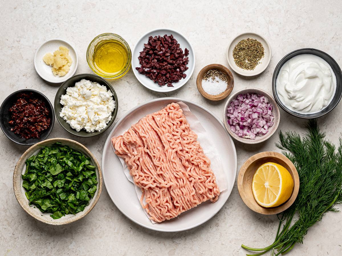 Ingredients for Greek chicken burgers and feta yogurt sauce arranged in individual bowls, including ground chicken, vegetables, herbs, feta, and yogurt.