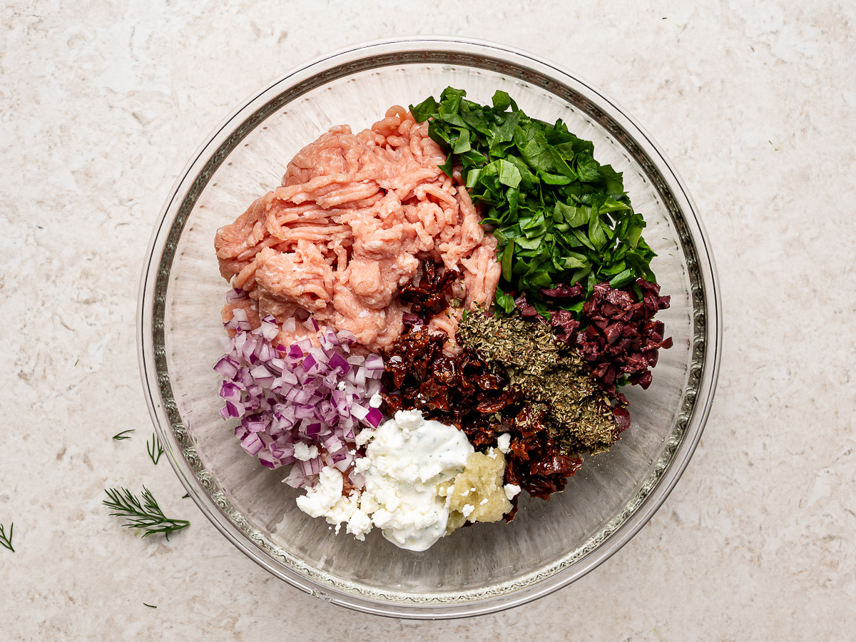 Ground chicken, vegetables, herbs, and seasonings being combined with feta yogurt sauce in a glass bowl.