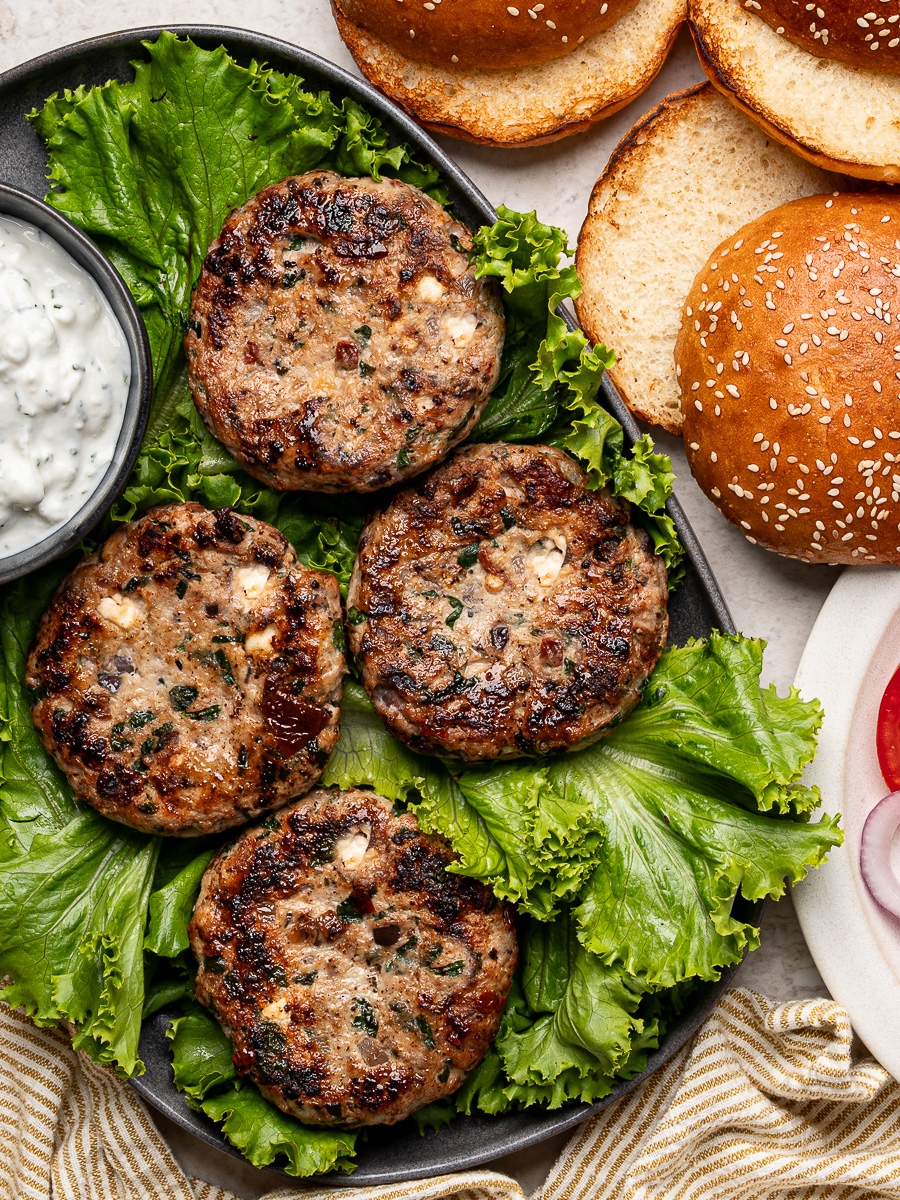 Four cooked chicken burger patties arranged on lettuce leaves on a platter, with burger toppings and feta yogurt sauce served alongside.