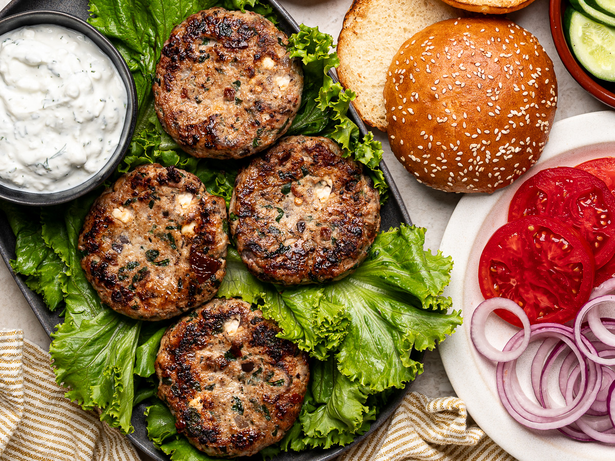Four cooked chicken burger patties arranged on lettuce leaves on a platter, with burger toppings and feta yogurt sauce served alongside.