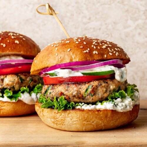 Two Greek chicken burgers on a wooden board, filled with feta yogurt sauce, lettuce, tomato, red onion, and cucumber.