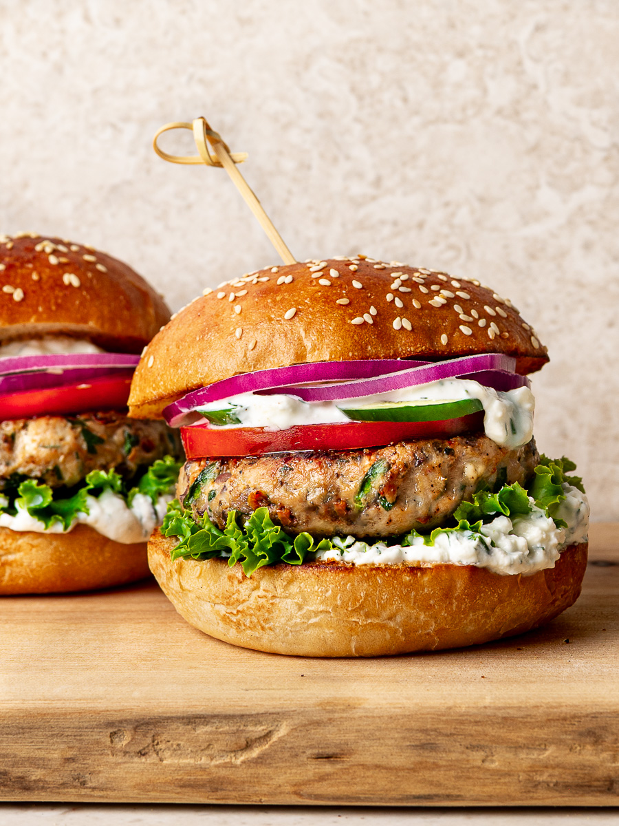 Two Greek chicken burgers on a wooden board, filled with feta yogurt sauce, lettuce, tomato, red onion, and cucumber.