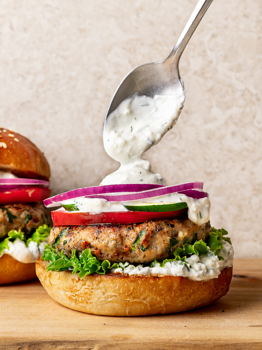 A spoonful of feta yogurt sauce is added to a cooked chicken burger patty on a bottom bun.