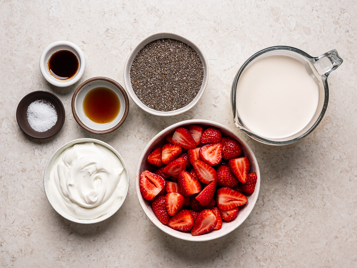 Ingredients for strawberries and cream chia pudding arranged in small bowls, including fresh strawberries, milk, Greek yogurt, chia seeds, maple syrup, vanilla, and kosher salt.