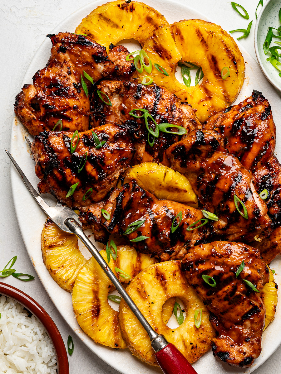 Huli Huli chicken thighs arranged on a platter with grilled pineapple rings, served with white rice in a bowl on the side.