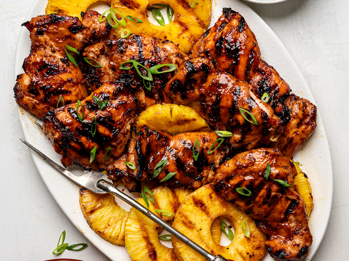 Close up of Huli Huli chicken thighs arranged on a white platter with grilled pineapple rings.