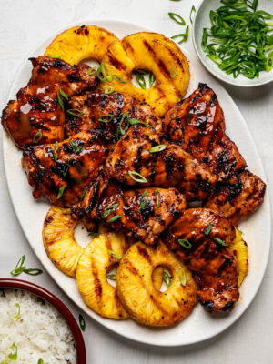 Huli Huli chicken thighs arranged on a white platter with grilled pineapple rings, served with white rice in a bowl on the side.