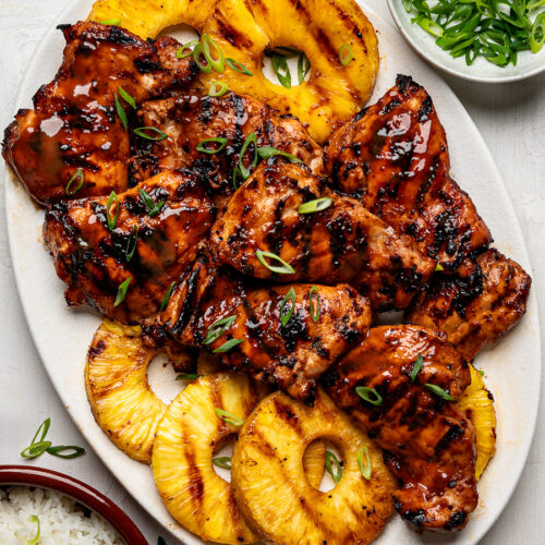 Huli Huli chicken thighs arranged on a white platter with grilled pineapple rings, served with white rice in a bowl on the side.