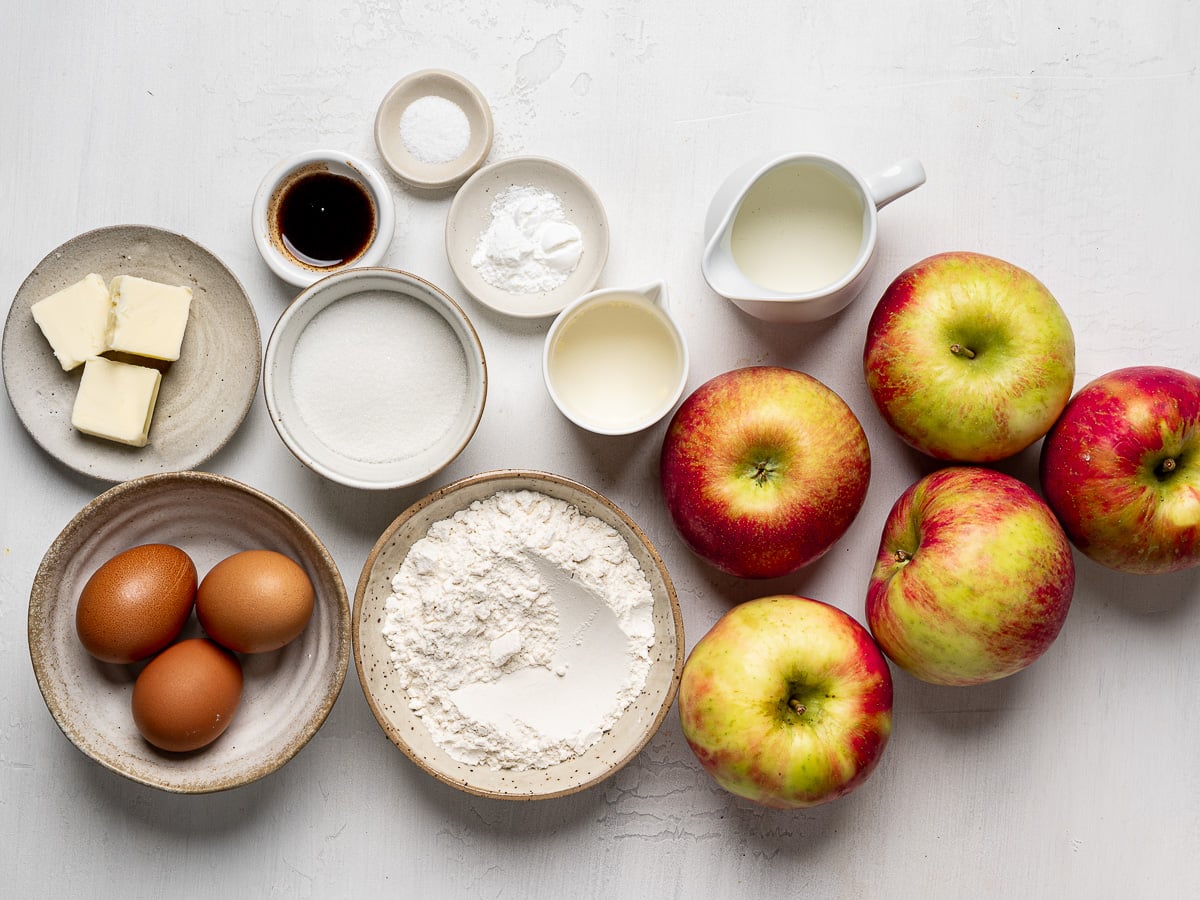 Ingredients for French apple cake such as eggs, butter, milk, sugar, baking soda, salt and whole apples prepared in bowls.